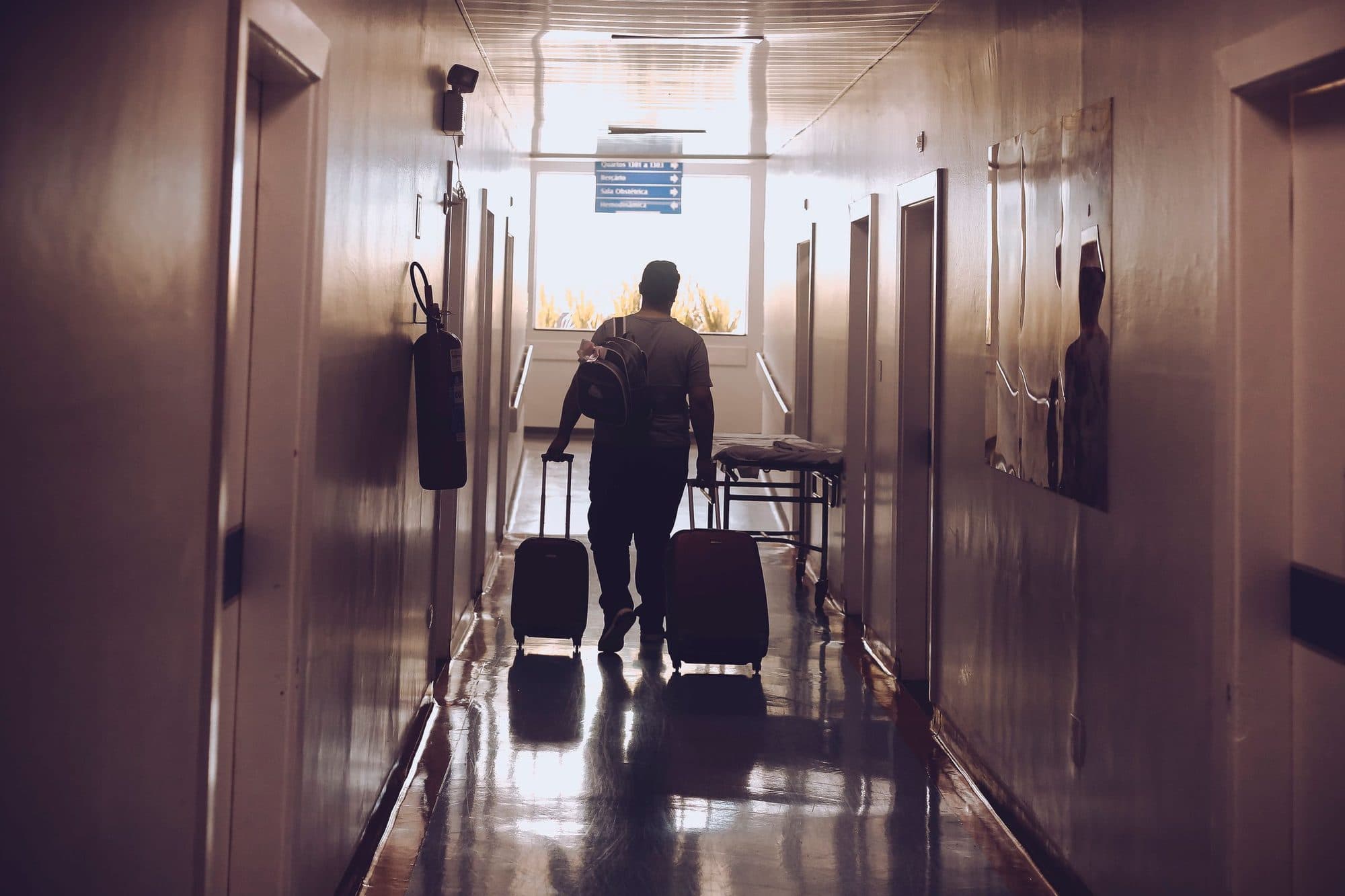 A guest with two suitcases walking down a hotel corridor, in silhouette.
